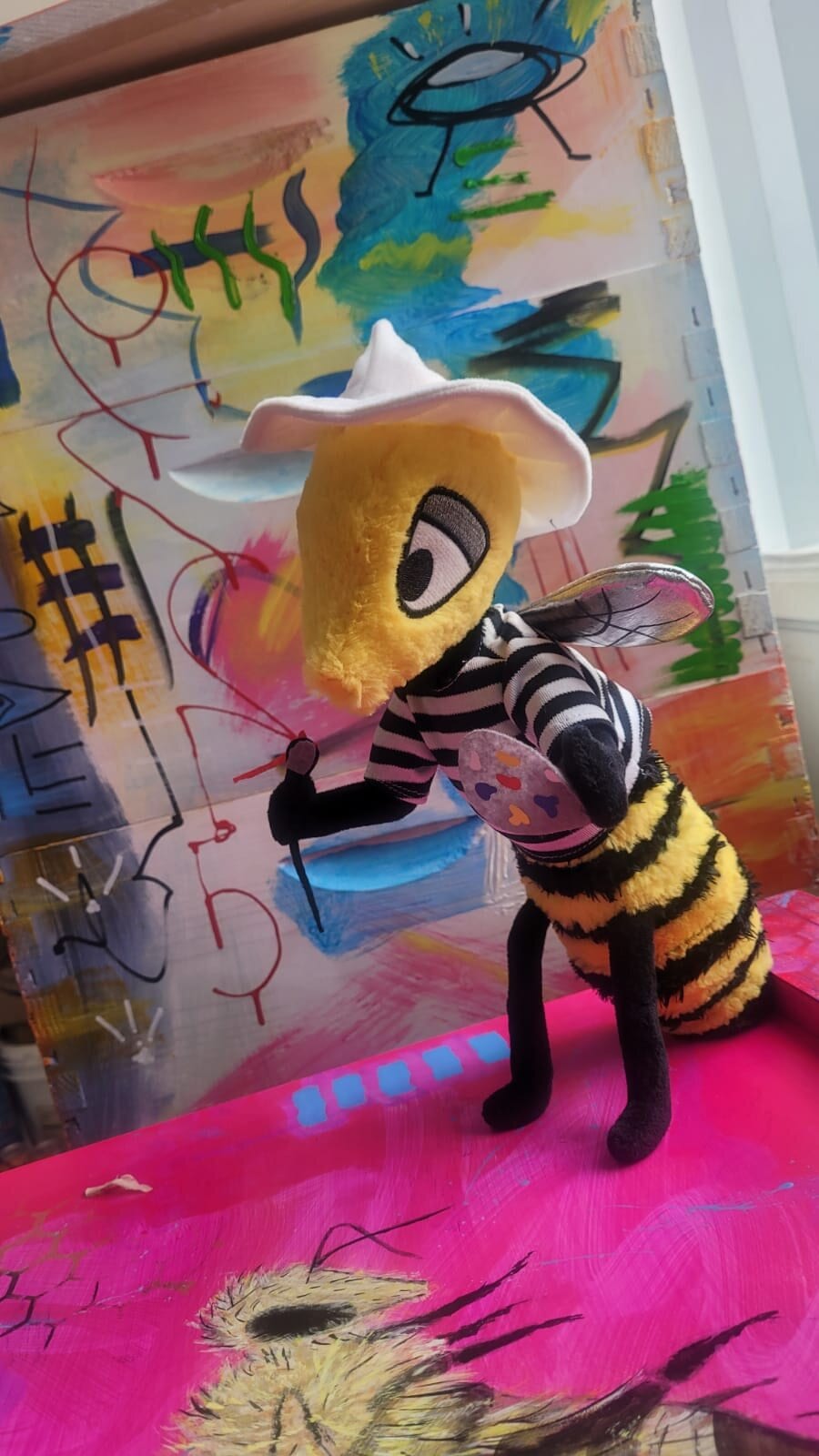 Beecasso's plush bee mascot perched at a paint-spattered easel