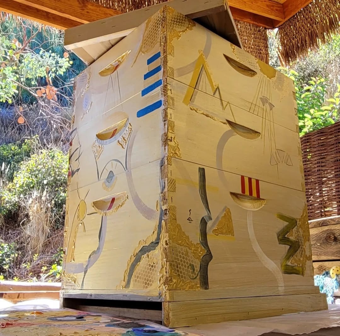 Beecasso painted bee box in cream and gold with geometric forms, photographed under thatched roof