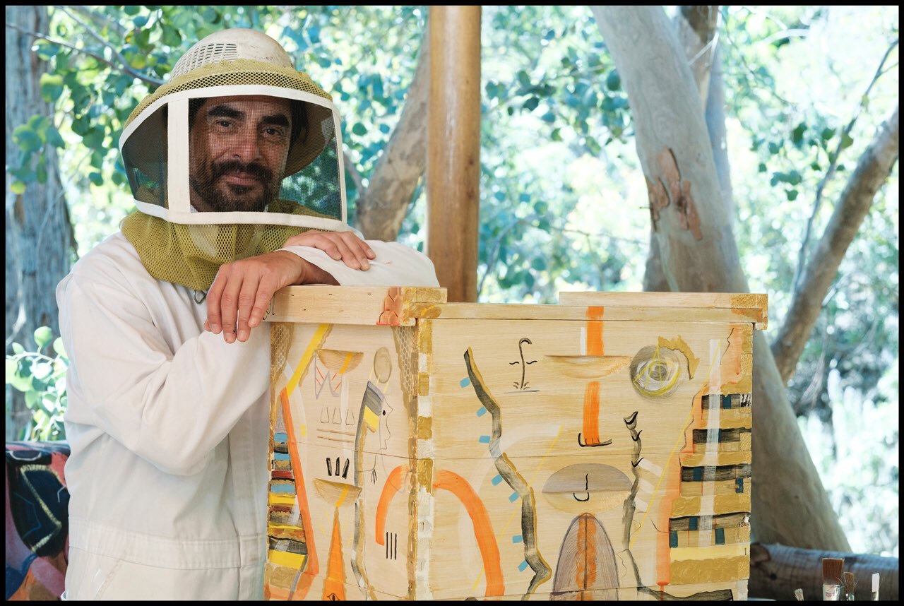 Steve Downs in beekeeper veil leaning on a painted Beecasso hive box in shaded outdoor setting