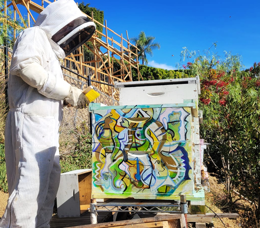 Steve Downs in bee suit inspecting a painted hive lid with colorful abstract art outdoors