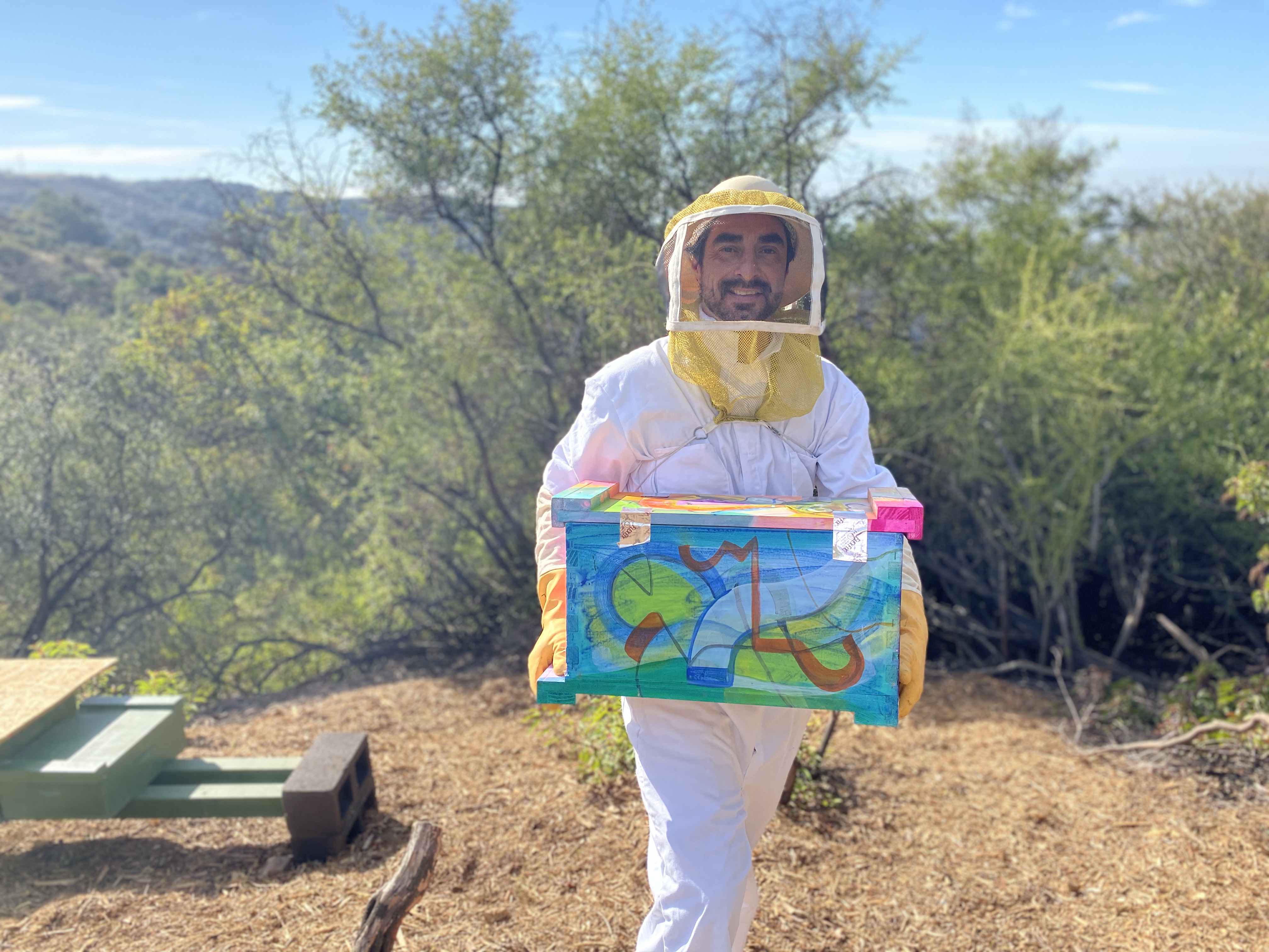 Steve Downs in bee suit carrying a painted Beecasso hive box through an outdoor hillside setting