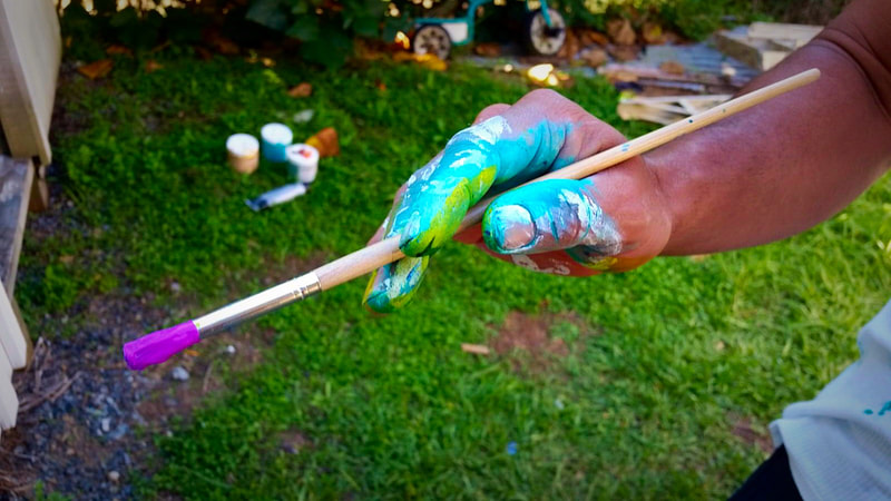 Close-up of Steve Downs' paint-covered hand gripping a brush with paints and grass visible in background
