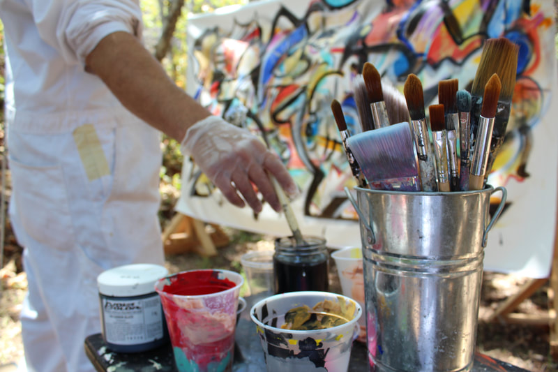Steve Downs at his outdoor studio in white suit, bucket of brushes in foreground, colorful canvas behind