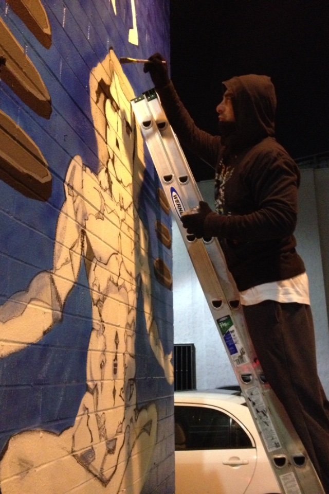 Steve Downs on a ladder painting the Hollywood mural at night, white figure emerging on blue wall