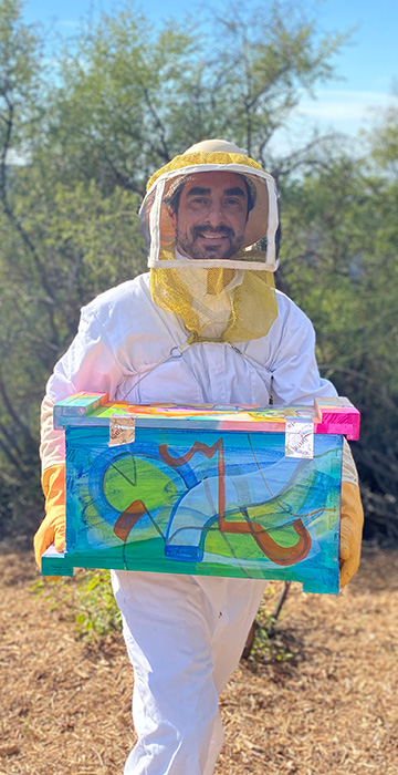 Steve Downs in full bee suit smiling while carrying a colorful painted Beecasso hive box outdoors